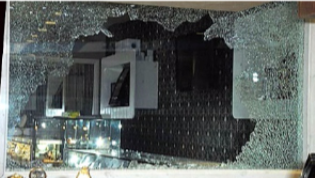 A large, shattered glass display window in a commercial interior with a dark, textured wall visible through the damage.
