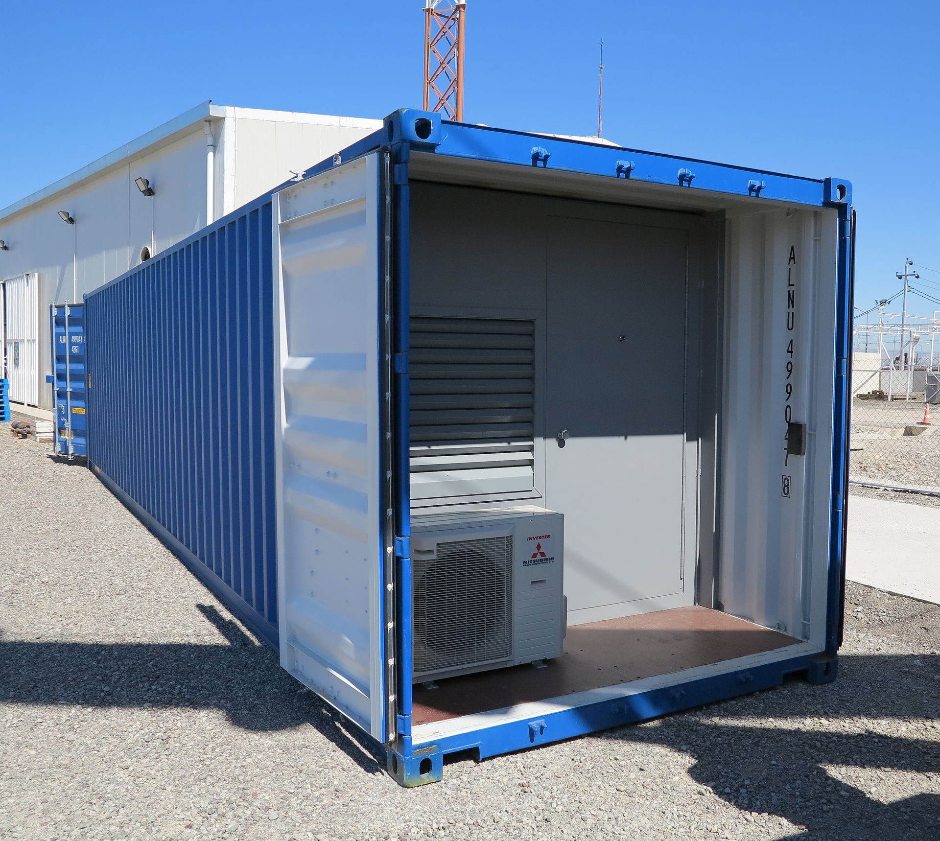 A blue and white shipping container sits on gravel, containing a grey wall and an installed air conditioning unit.