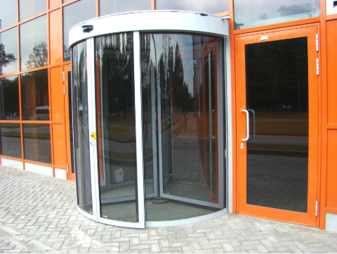 A glass revolving door entry next to a standard orange-framed glass door on a building exterior with brick paving.