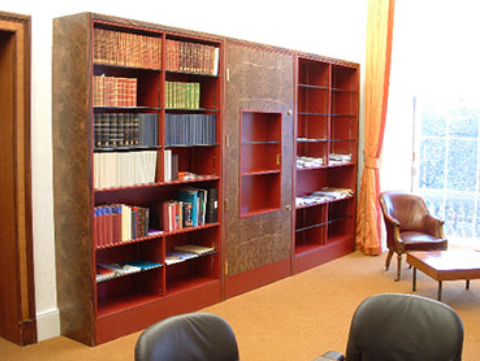 A large, reddish-brown wooden bookcase spanning a wall in an office, with two armchairs in the foreground.