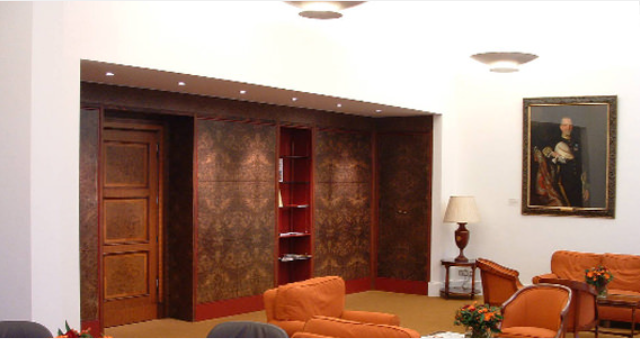 A formal lounge features brown patterned walls, a wooden door, orange chairs, and a portrait hanging on a white wall.