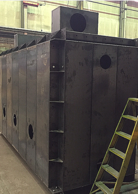 A large, dark metal industrial tank with circular ports, reinforced corner beams, and a yellow stepladder in a workshop.
