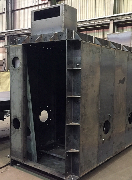 A large, rectangular steel industrial structure with cutouts and support brackets, standing in a workshop.