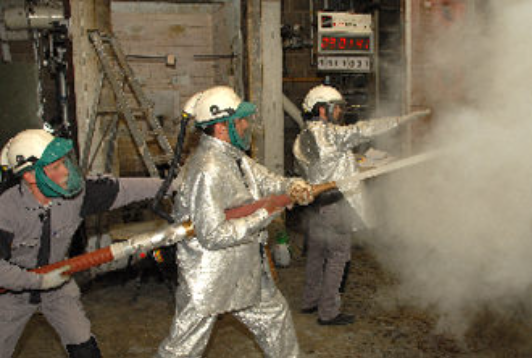 Three firefighters in protective silver gear and helmets spraying a hose into a cloud of white smoke inside a room.