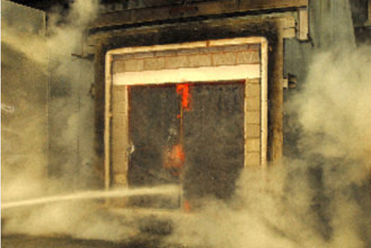 A jet of water from a fire hose hits a dark, smoky doorway framed by concrete blocks during a firefighting exercise.