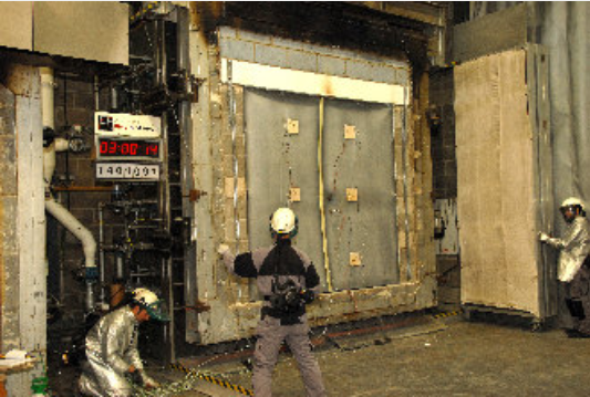 Technicians in protective suits monitor a fire-resistance test on a large panel at a laboratory facility.