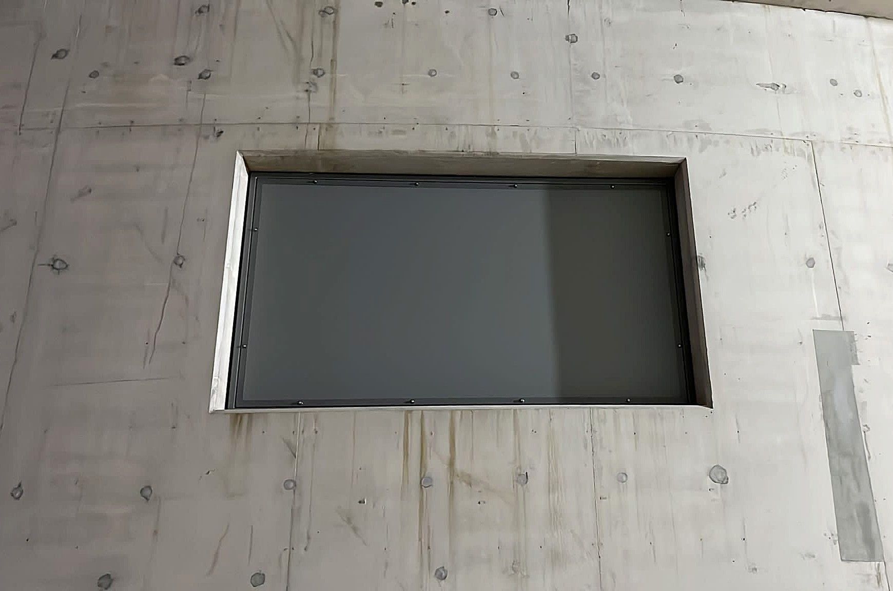 A rectangular window framed in raw concrete, showing a dark, reflective surface within a light gray wall.