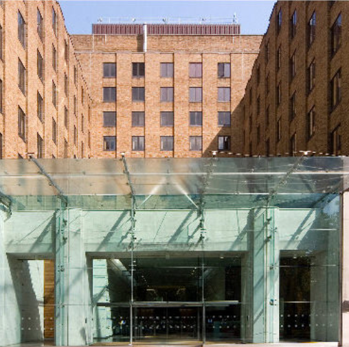 The entrance of a tan brick building with a modern glass canopy over the glass doors.