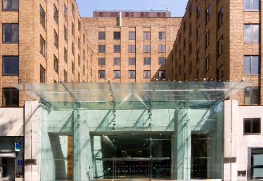 A modern glass canopy entrance attached to a multi-story brown brick building with a symmetrical facade.