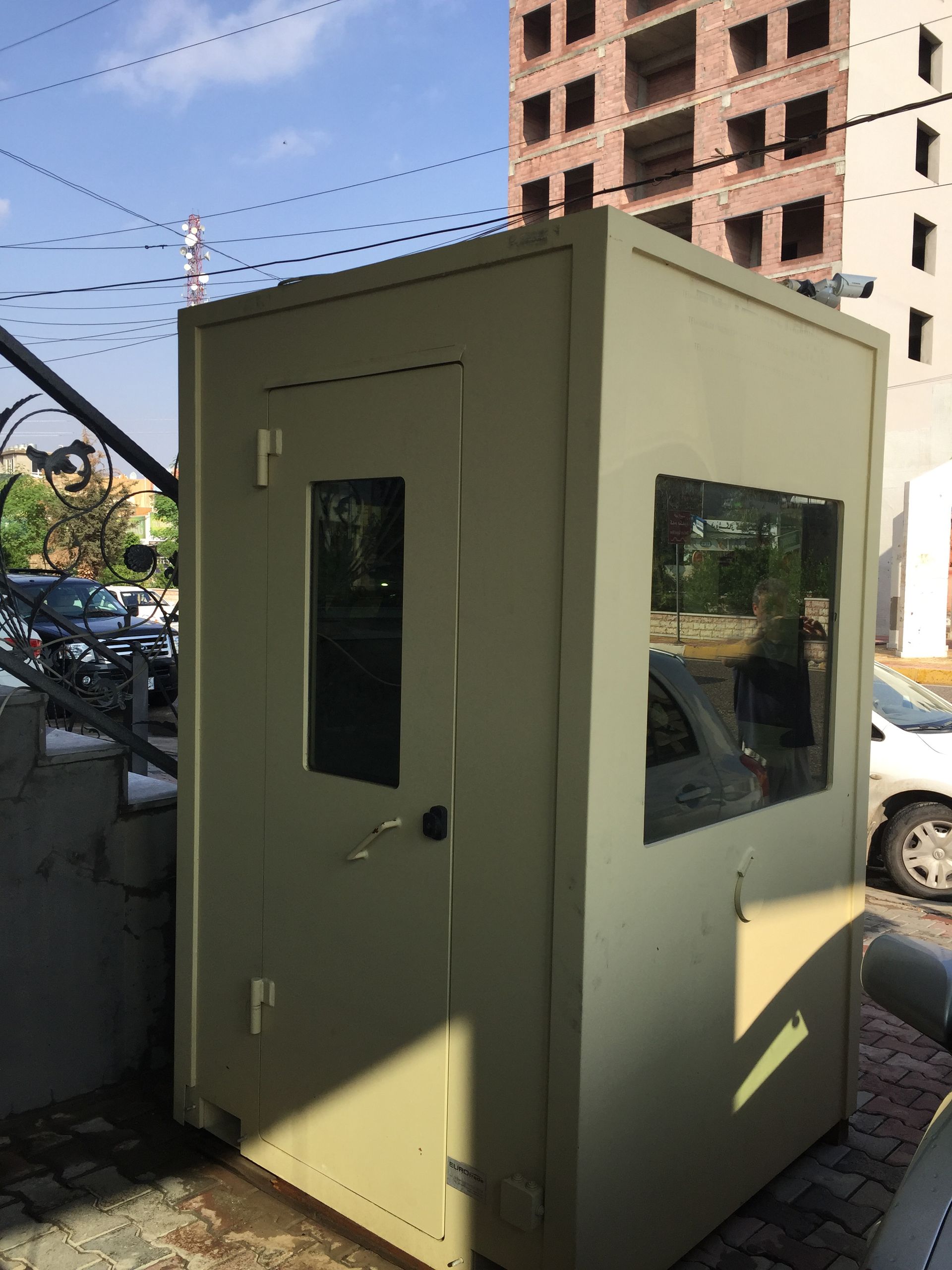 A beige portable security guard booth with a door and windows, situated outdoors near a building and parked cars.