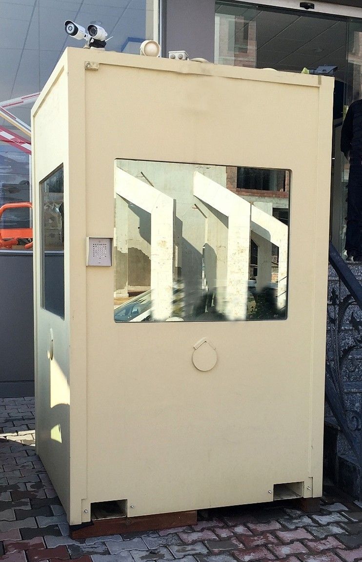 A cream-colored metal security booth with a surveillance camera mounted on the roof sits on a paved surface.