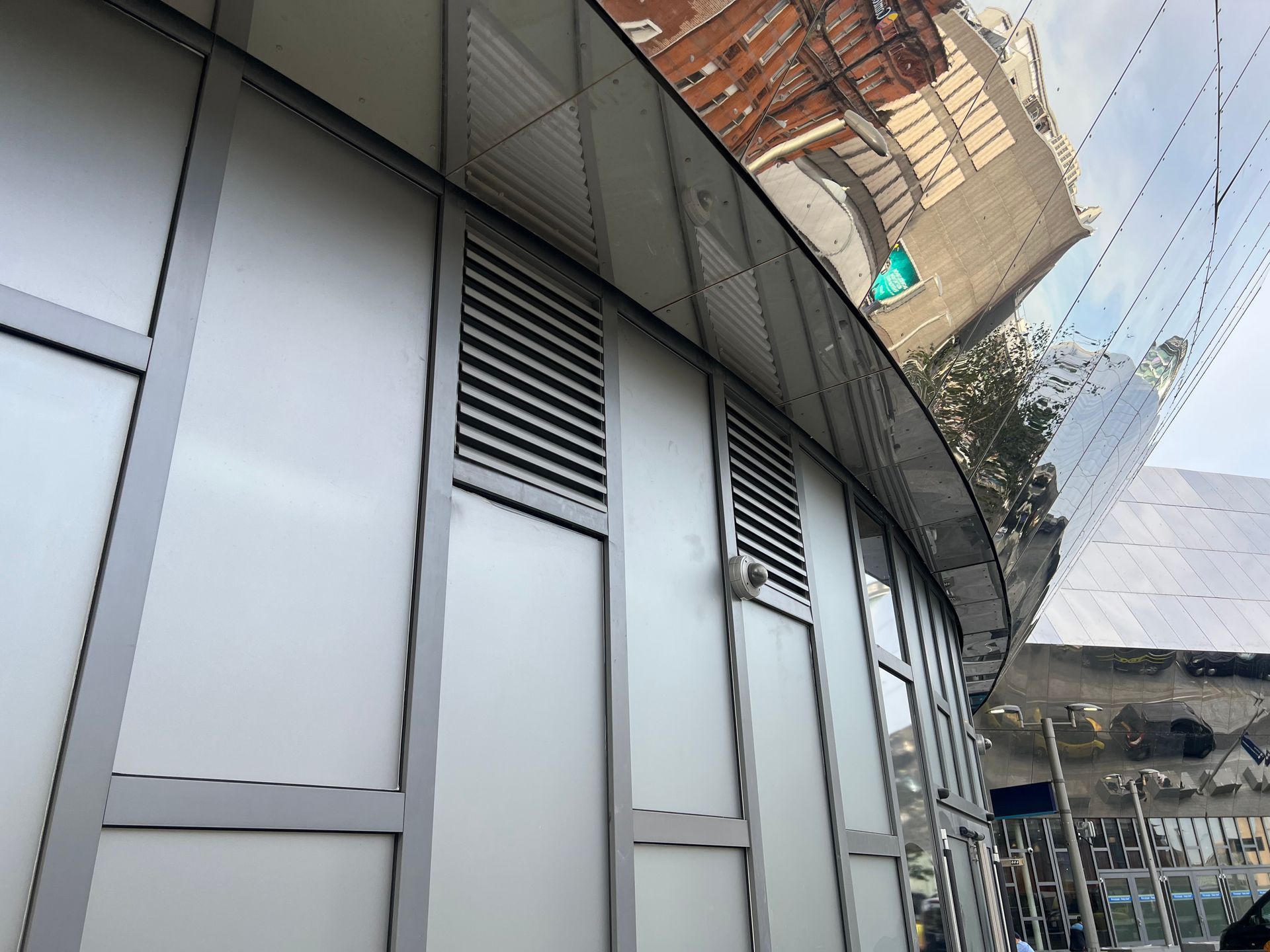 A low-angle view of a curved building facade featuring light grey panels, metal trim, and rectangular ventilation grilles.