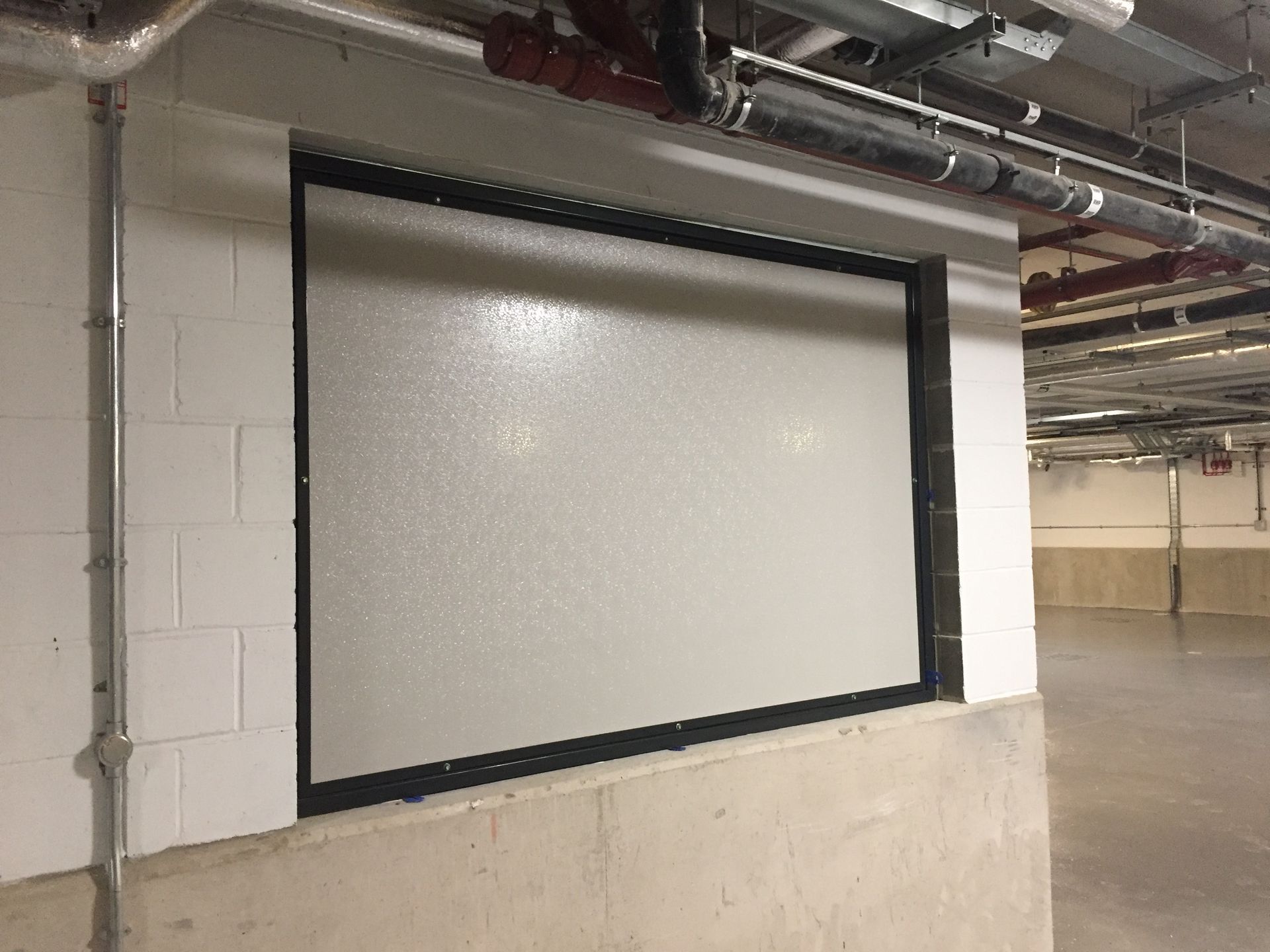 A rectangular, light-gray panel with a black frame mounted on a white cinder block wall in a parking garage.