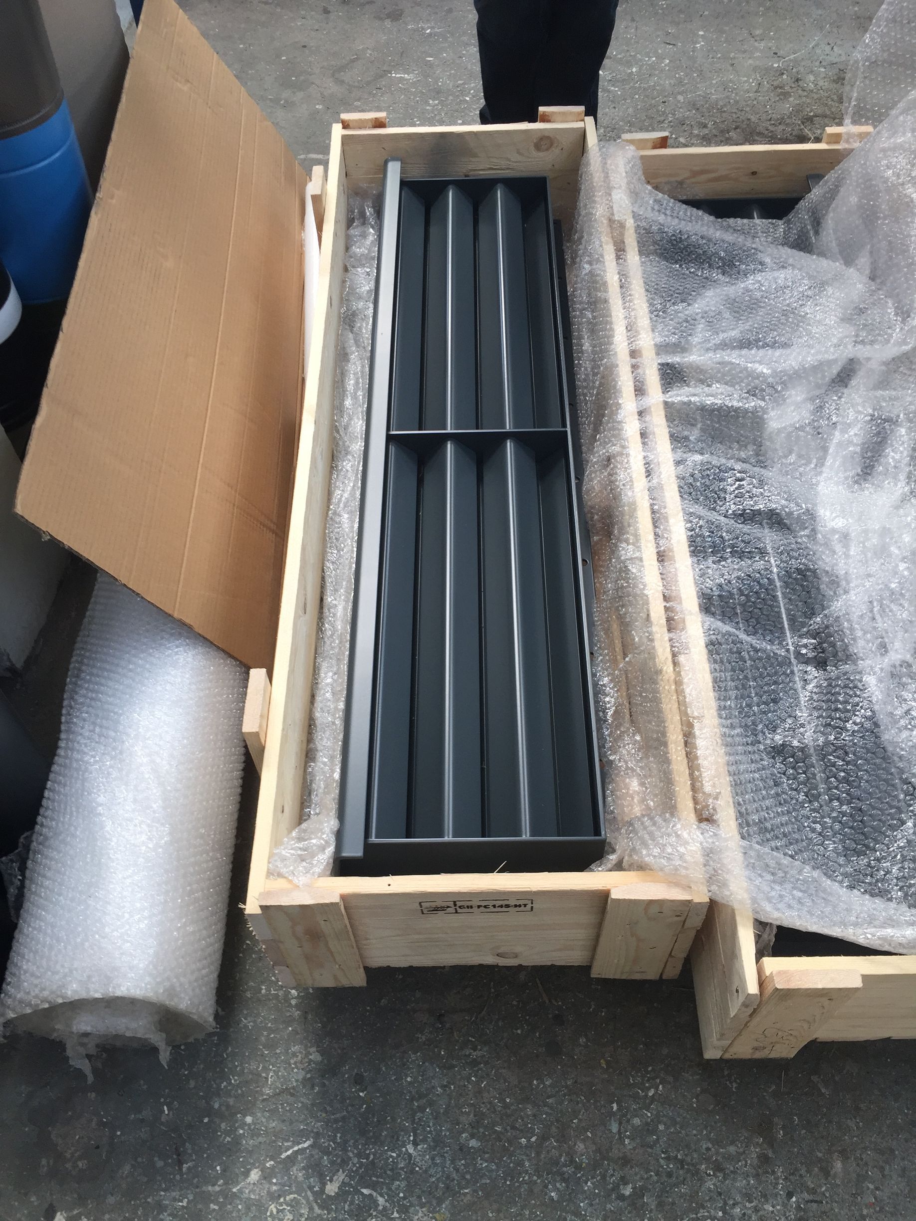 A black metal louver panel packed in a wooden shipping crate, surrounded by bubble wrap on a concrete surface.
