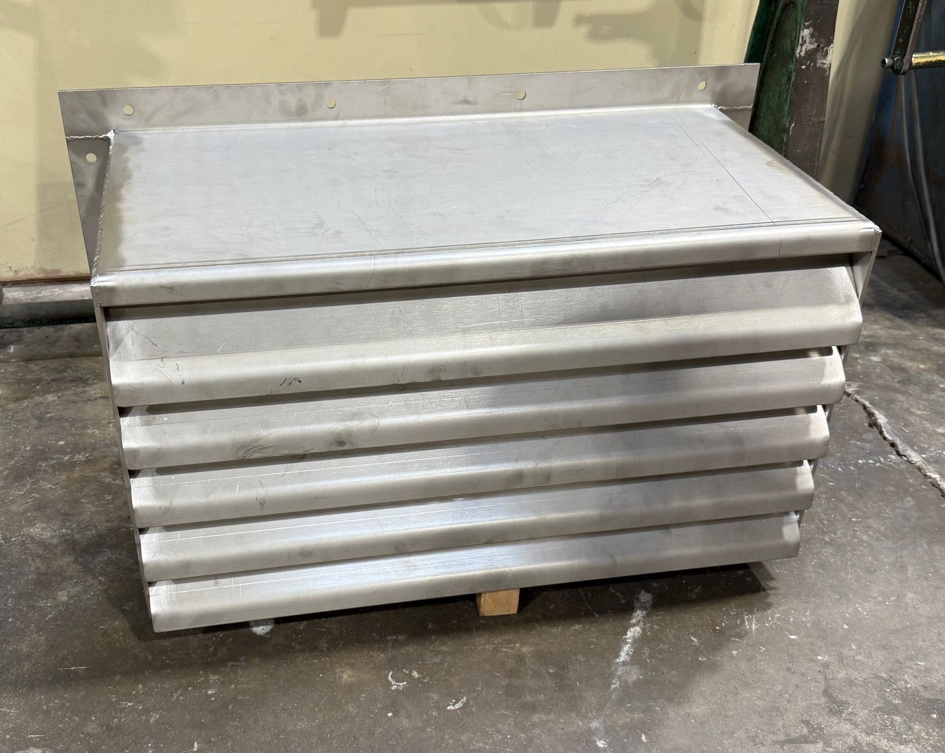 A metal industrial louvered vent unit with horizontal slats, resting on a wooden block on a concrete floor.