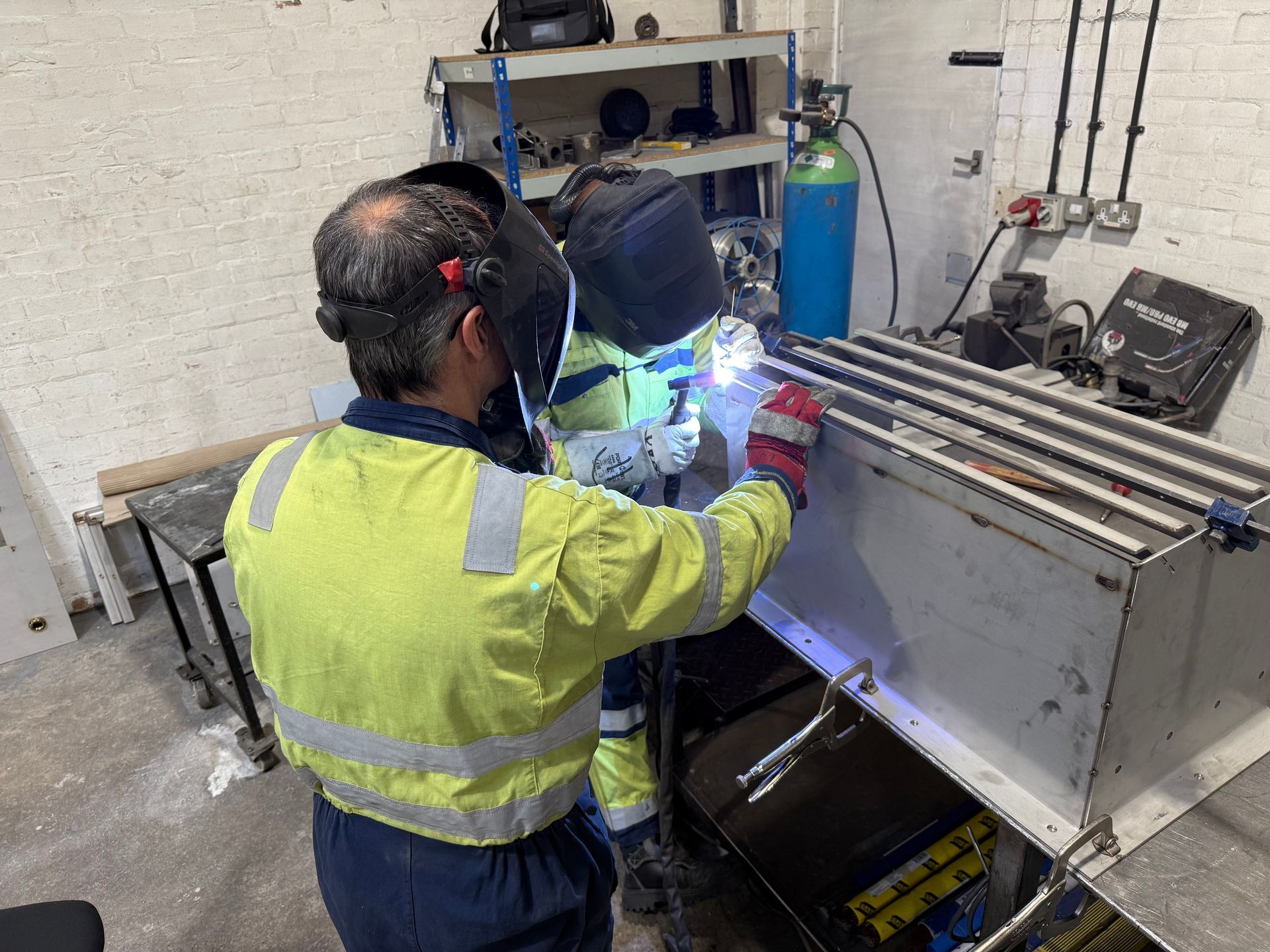 Two people in high-visibility work gear and welding helmets weld a metal component at a workshop bench.