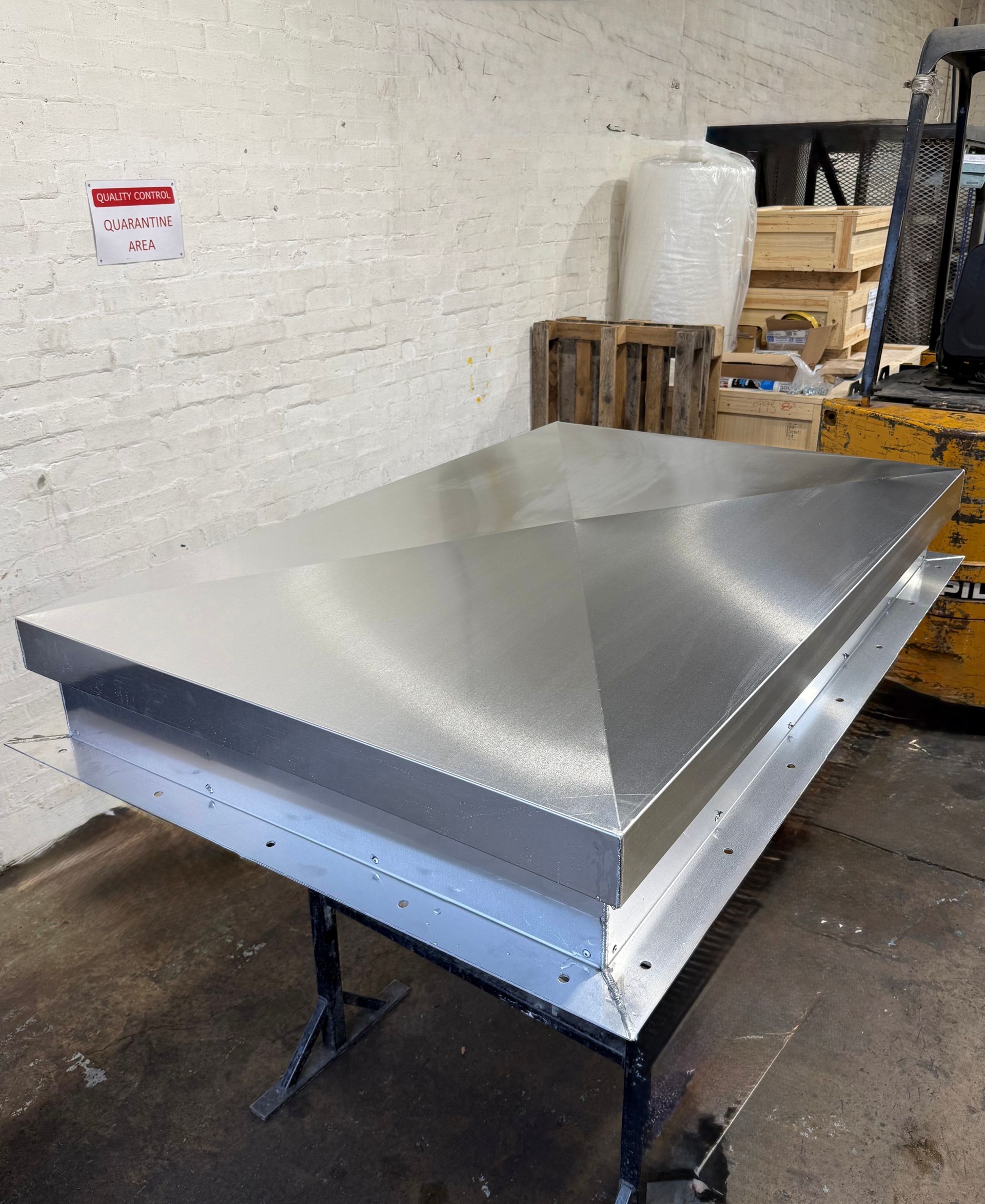 A rectangular, metallic roof vent cap with a peaked top sits on a metal stand inside a workshop.