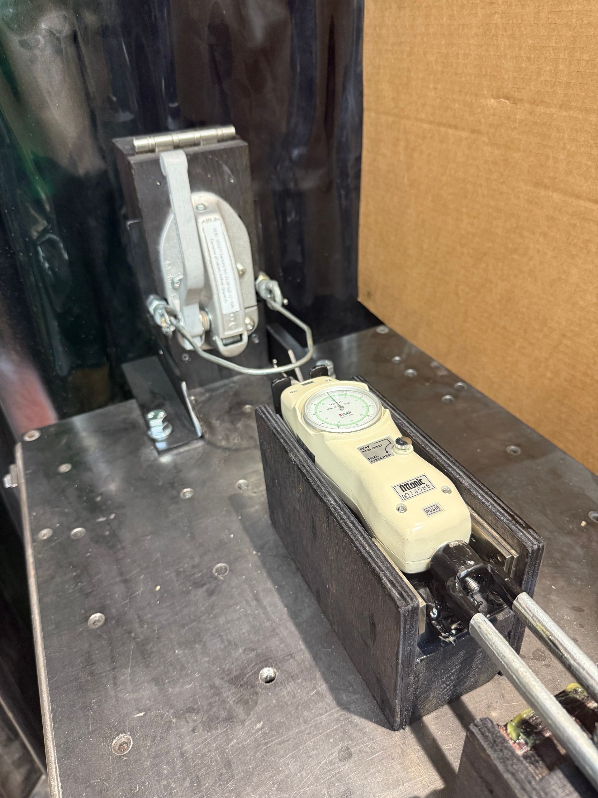 A handheld force gauge mounted in a metal fixture on a workstation, connected to a tension cable and a lever mechanism.