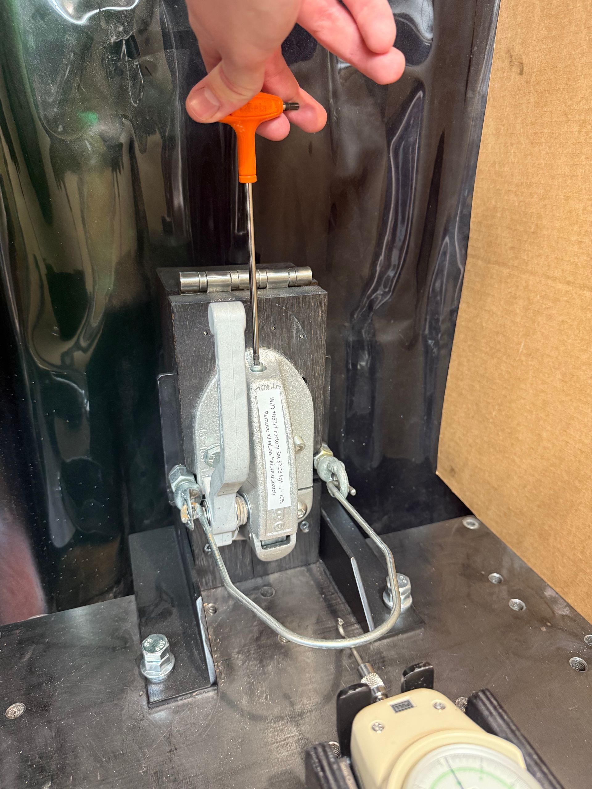 A hand uses an orange T-handle tool to adjust a mechanical locking latch mounted on a metallic surface.
