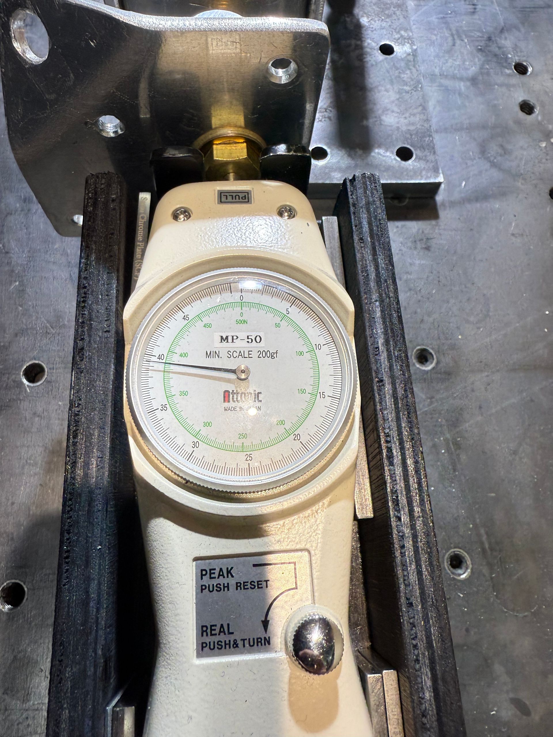 An analog force gauge mounted on a dark metal workspace, measuring force with its needle pointing left on a white dial.
