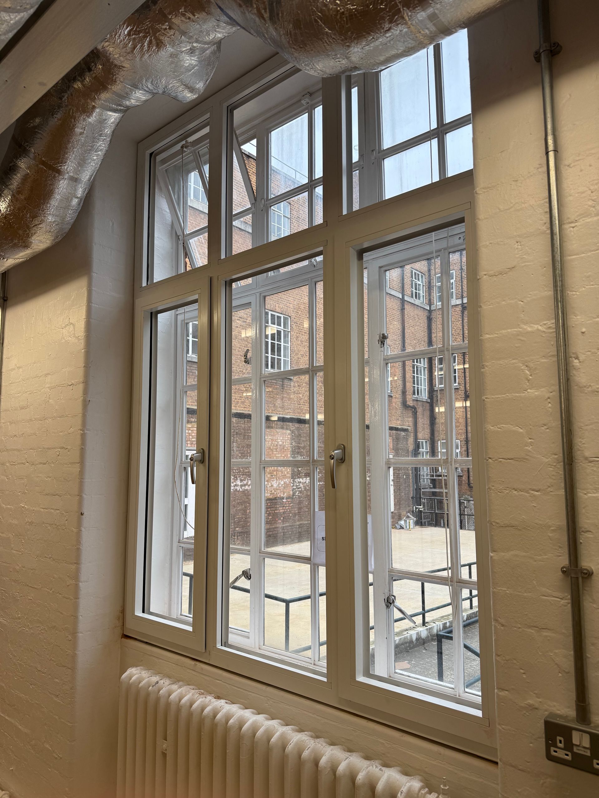 A white-framed, multi-pane window overlooks a brick building and outdoor walkway, with a radiator visible below.