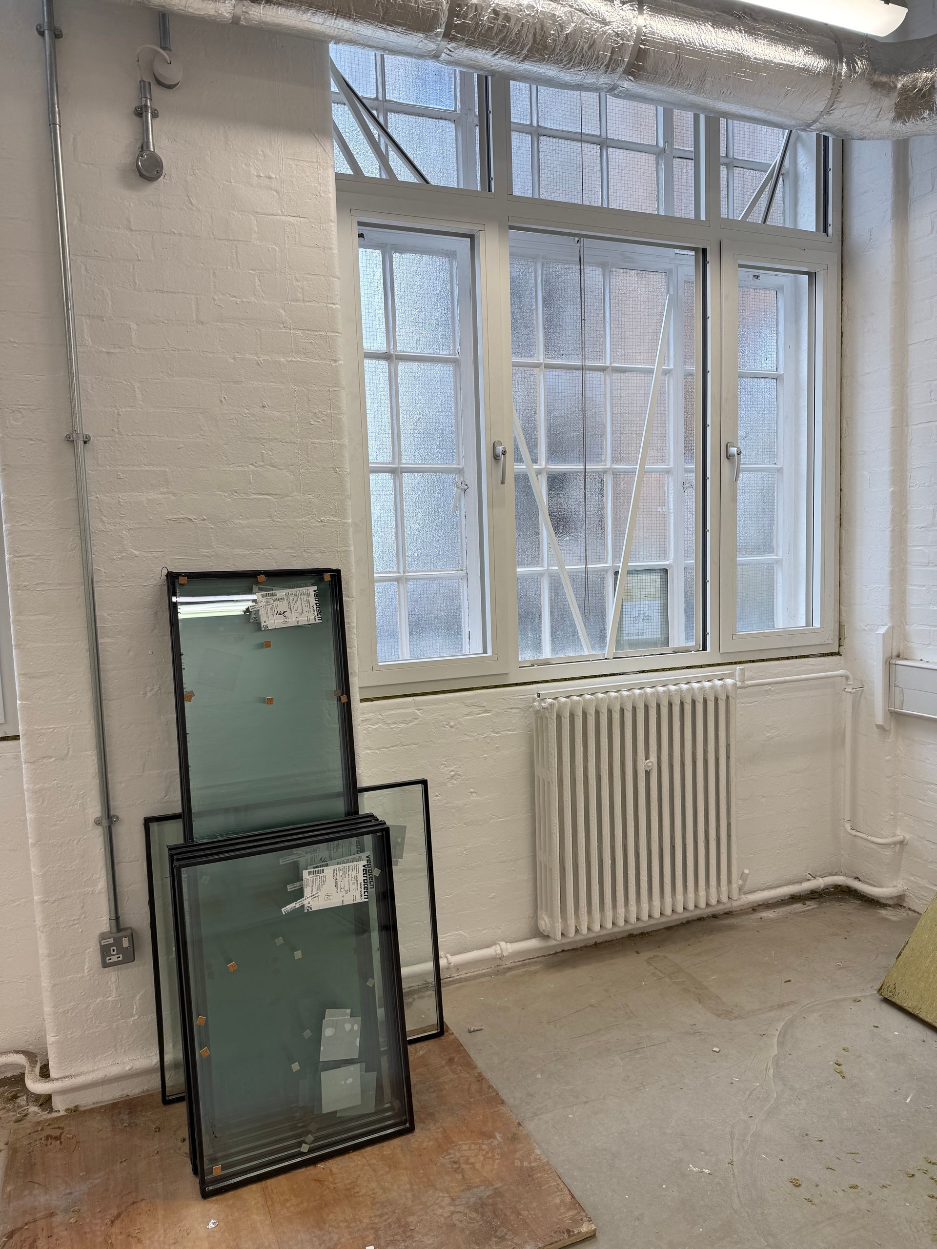 Two glass window panels lean against a white, textured wall near a large window and radiator in an unfinished room.