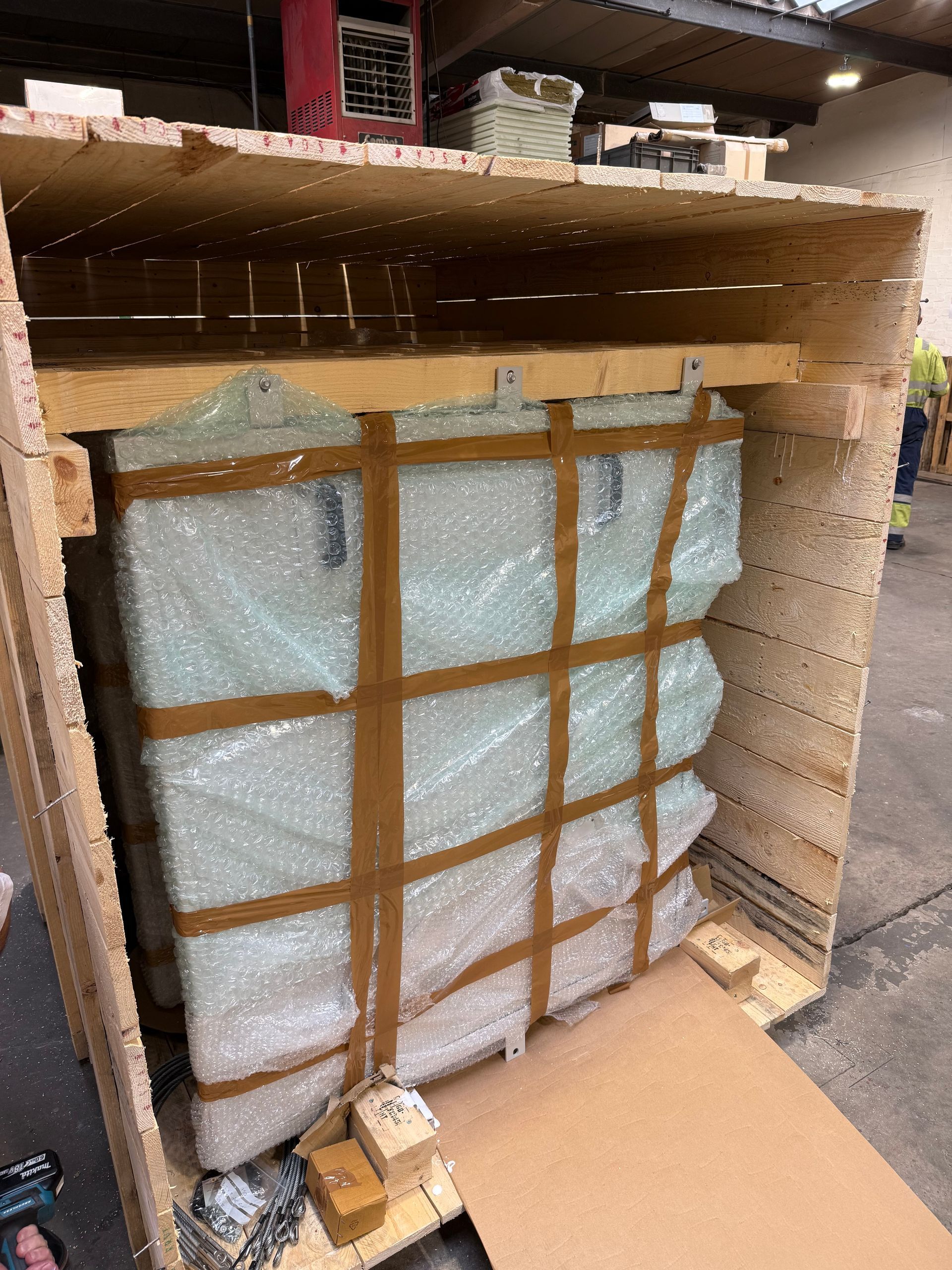 A large, bubble-wrapped item secured with brown tape inside a wooden shipping crate on a warehouse floor.