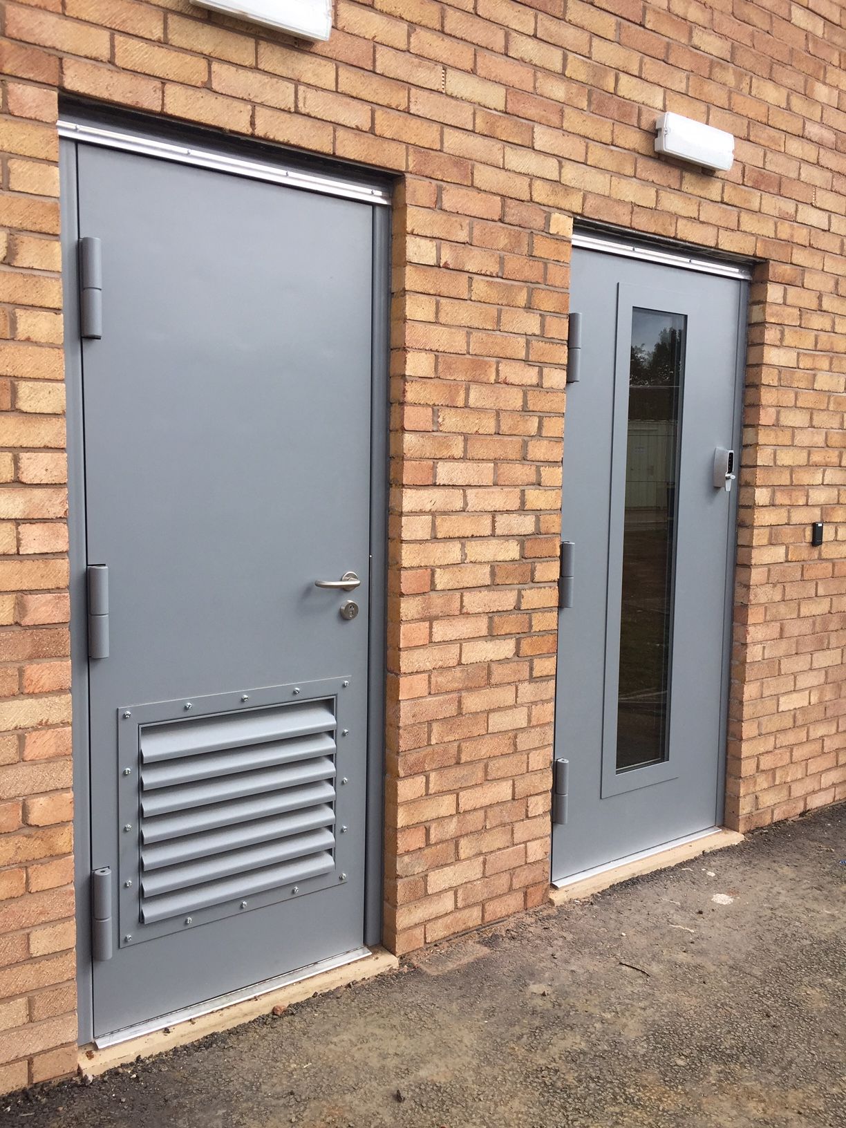 Two grey metal doors set in a tan brick wall; the left door has a ventilation grate, and the right door has a vertical window.