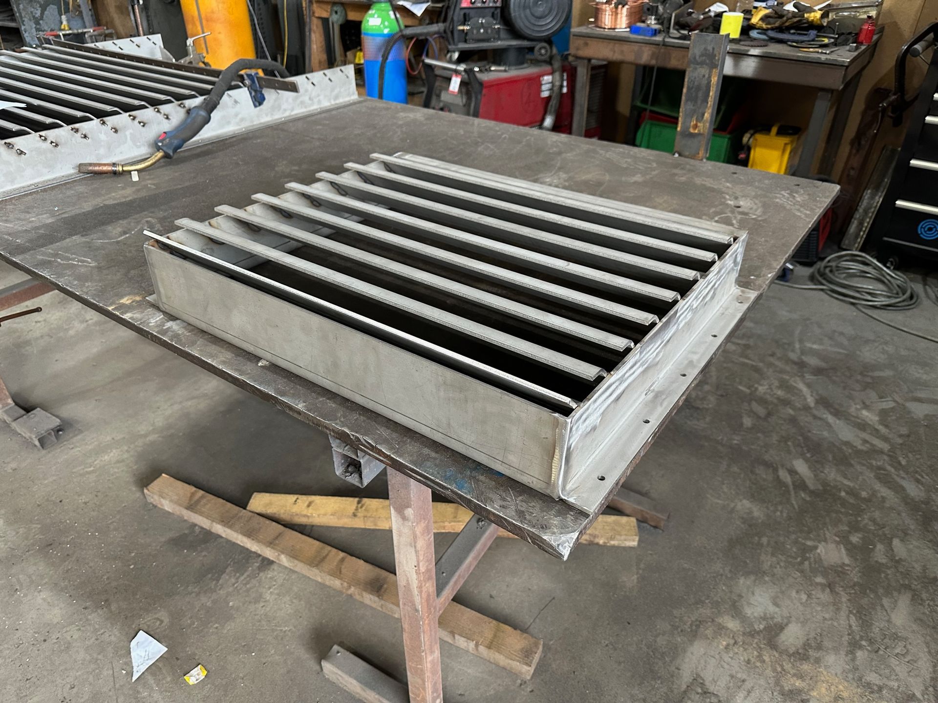 A square, metal slatted vent or grate sits on a workshop table in a fabrication shop.