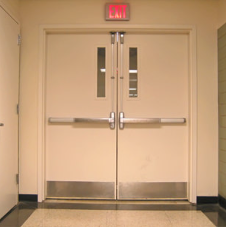 Double beige doors with crash bars and small windows, topped with a red illuminated EXIT sign in a hallway.