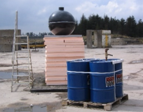 A spherical black object sits atop a stack of light-colored foam blocks next to blue drums on a wooden pallet.