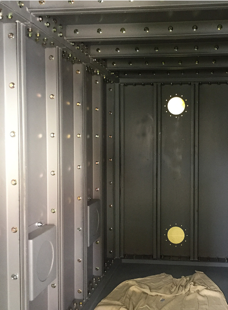 An interior view of a gray metal container featuring riveted wall panels, structural framing, and two circular portholes.