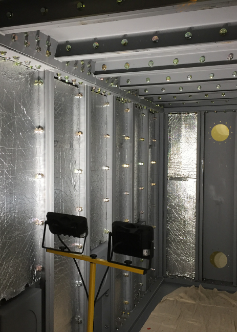 A portable work light sits in a metal, industrial enclosure under construction, featuring reflective insulation panels.