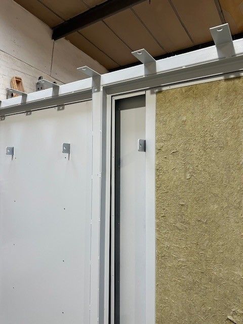 Construction site interior showing wall panels with metallic support brackets, insulation, and framing in a room.