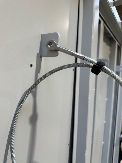 A metal cable is secured to a white wall using a small, silver mounting bracket and a screw.