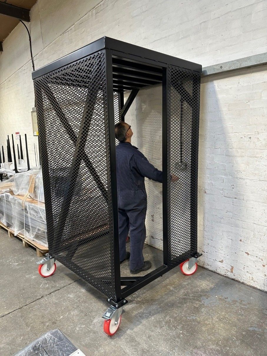 A person stands inside a tall, black metal cage on wheels with mesh sides in an industrial workshop.