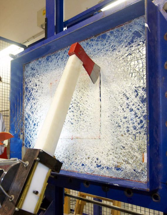 An axe mounted on a mechanical arm strikes a pane of shattered, reinforced security glass within a blue metal frame.