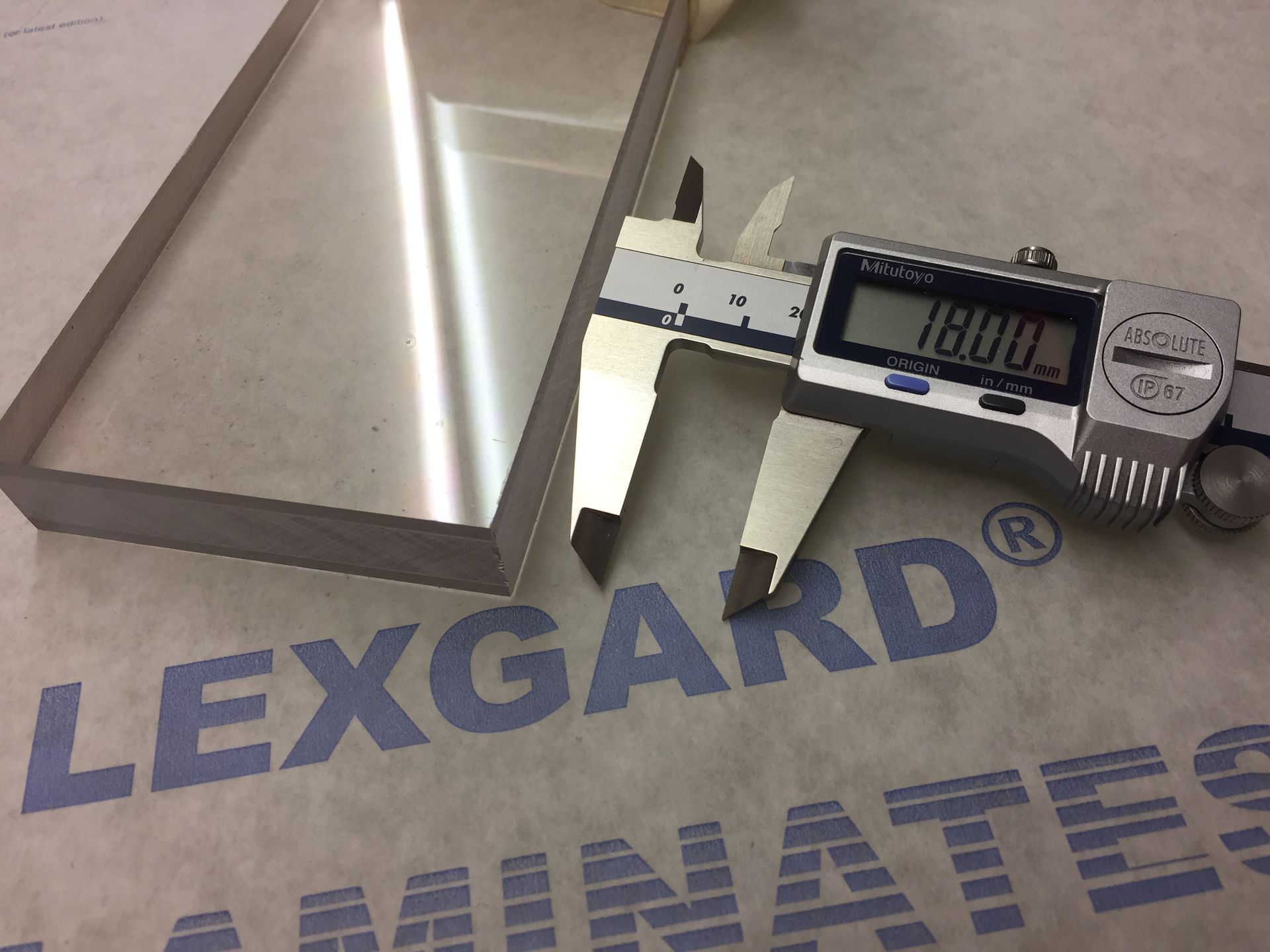A digital caliper measuring the thickness of a transparent Lexgard laminate sheet, showing a reading of 18.00 mm.