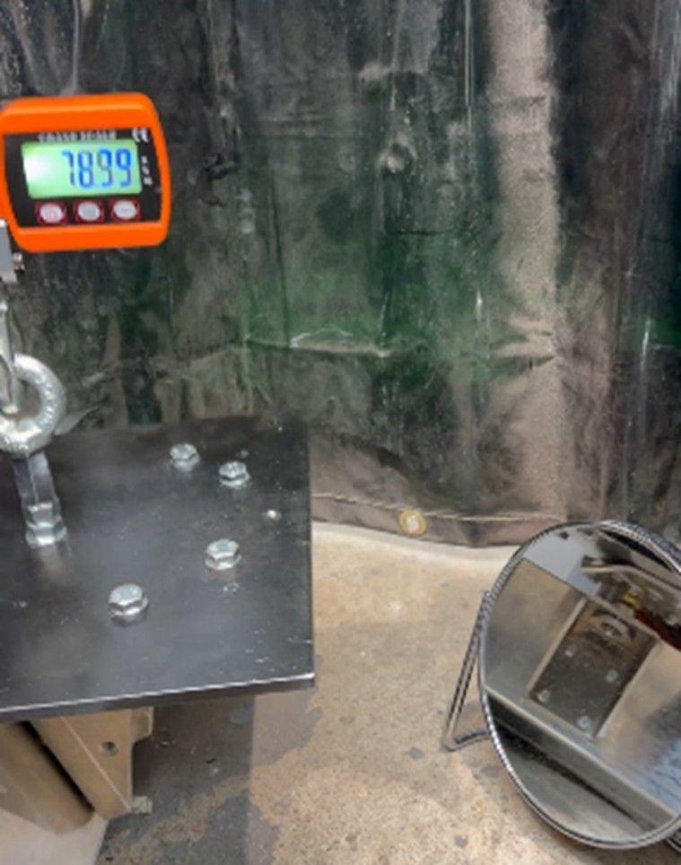 An orange digital hanging scale shows a reading of 7899, suspended above a metal plate with bolts near a reflective mirror.