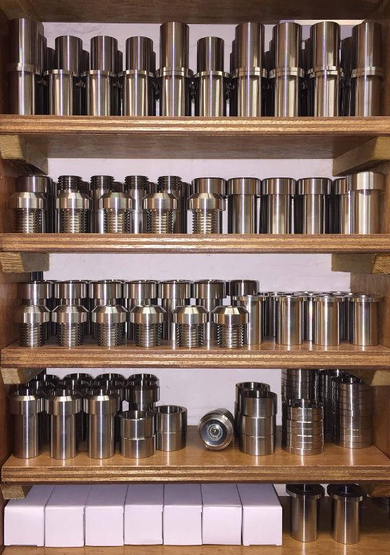 A wooden shelf unit holding rows of organized, metallic cylindrical parts and white rectangular boxes.