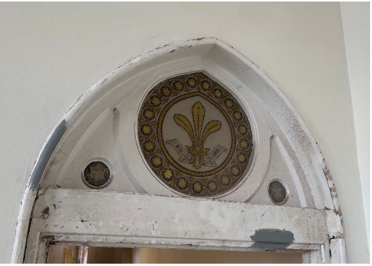 A pointed white arched doorway framing a central circular decorative emblem featuring a gold fleur-de-lis on a dark border.