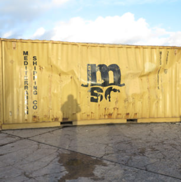 A dented, yellow MSC Mediterranean Shipping Co. container sits on cracked pavement under a cloudy blue sky.