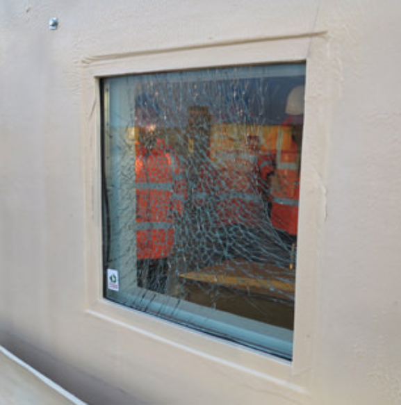 A shattered, tempered glass window pane in a beige wall, reflecting two people in high-visibility orange work vests.