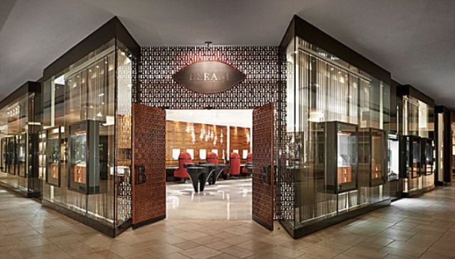 The storefront of an upscale jewelry shop with glass windows, warm interior lighting, and ornate wooden door panels.