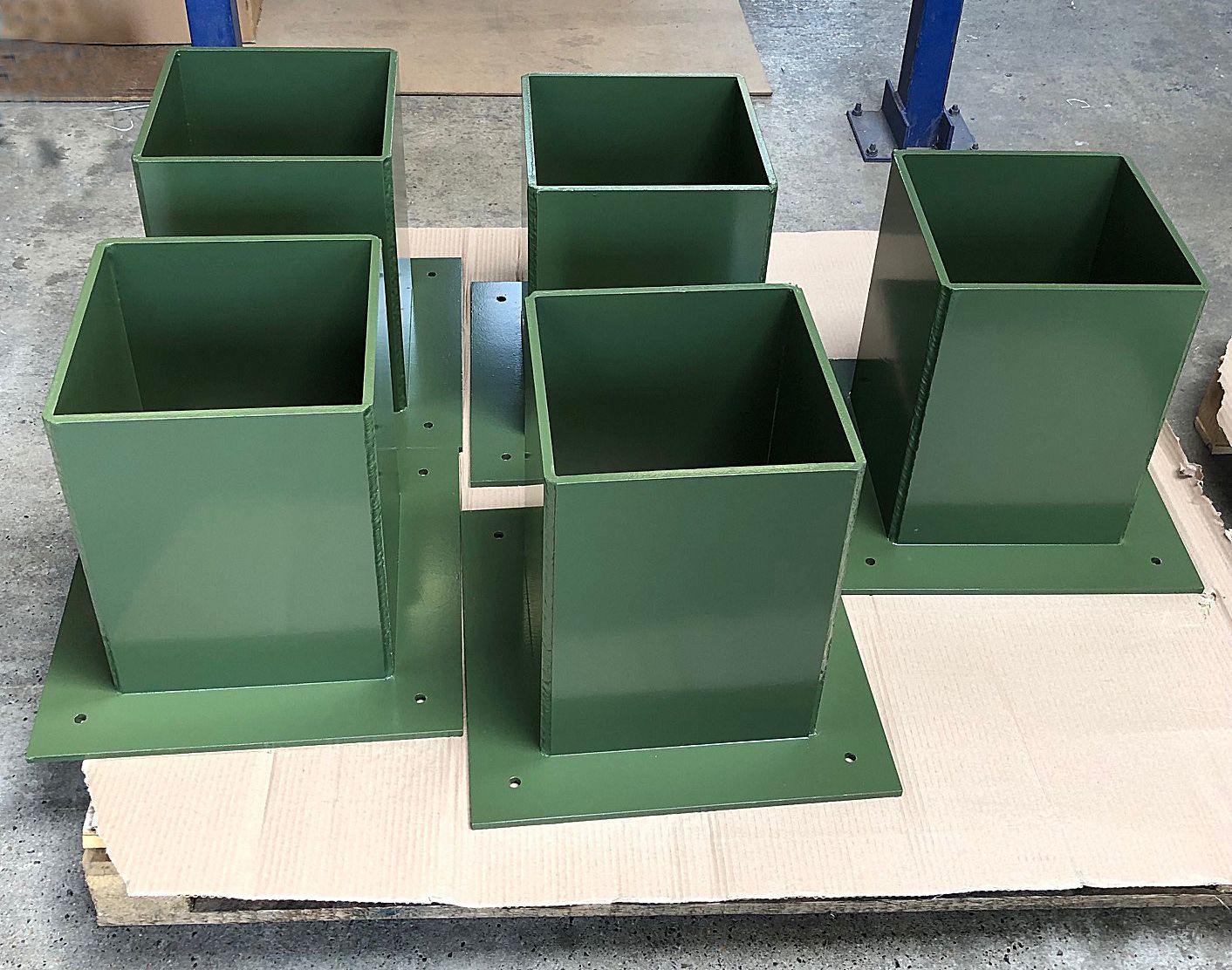 Five green, square-shaped steel post bases with mounting plates arranged on a cardboard-covered pallet.