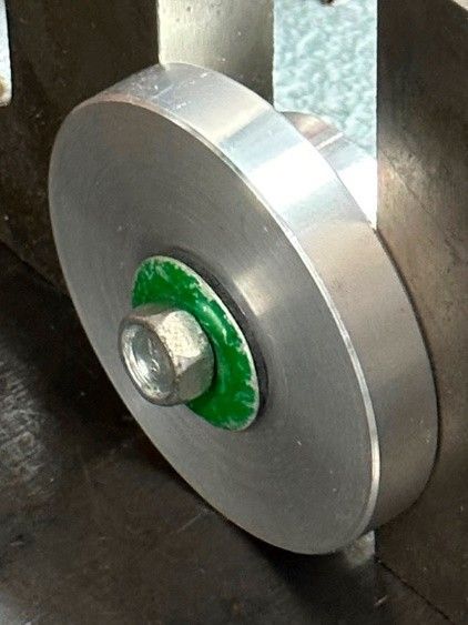 A silver metal circular component held in place by a bolt with a green washer against a dark industrial surface.