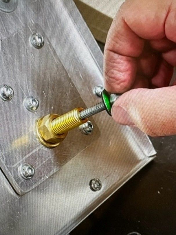 A hand holds a screw with a green washer, preparing to insert it into a brass fitting on a metallic panel.
