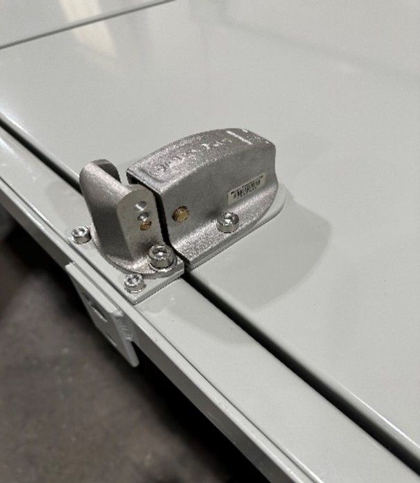 A metal latch mechanism mounted on a gray surface, featuring a hinged block secured by several screws.
