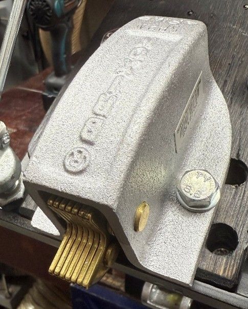 A silver metal belt sander tracking mechanism with a ribbed brass-colored tensioner, mounted on a workshop table.