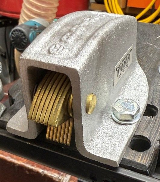 A silver metal cam cleat for securing rope, mounted on a dark workbench with a visible bolt.